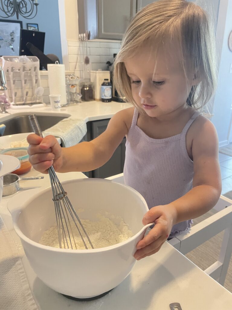A toddler girl in the kitchen stirring a bowl of flour making Hidden veggie pizza muffins perfect for toddlers topped with cheese and pepperoni.