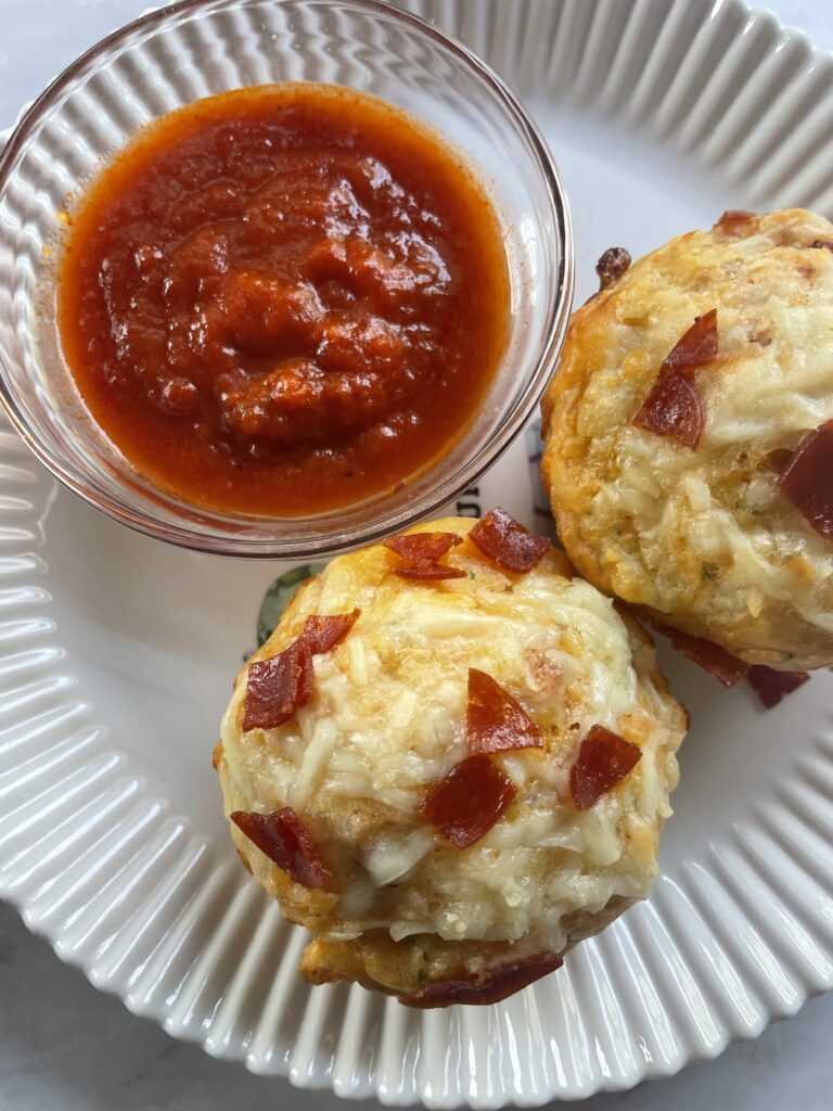 Hidden veggie pizza muffins perfect for toddlers topped with cheese and pepperoni sitting on a white plate with a side cup of tomato sauce for dipping.